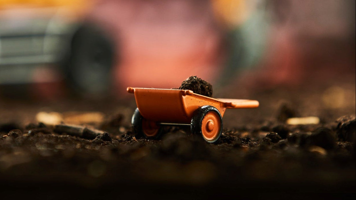 A miniature orange toy wheelbarrow filled with dark soil sits in sharp focus on dark dirt, with a blurred yellow construction vehicle in the background.